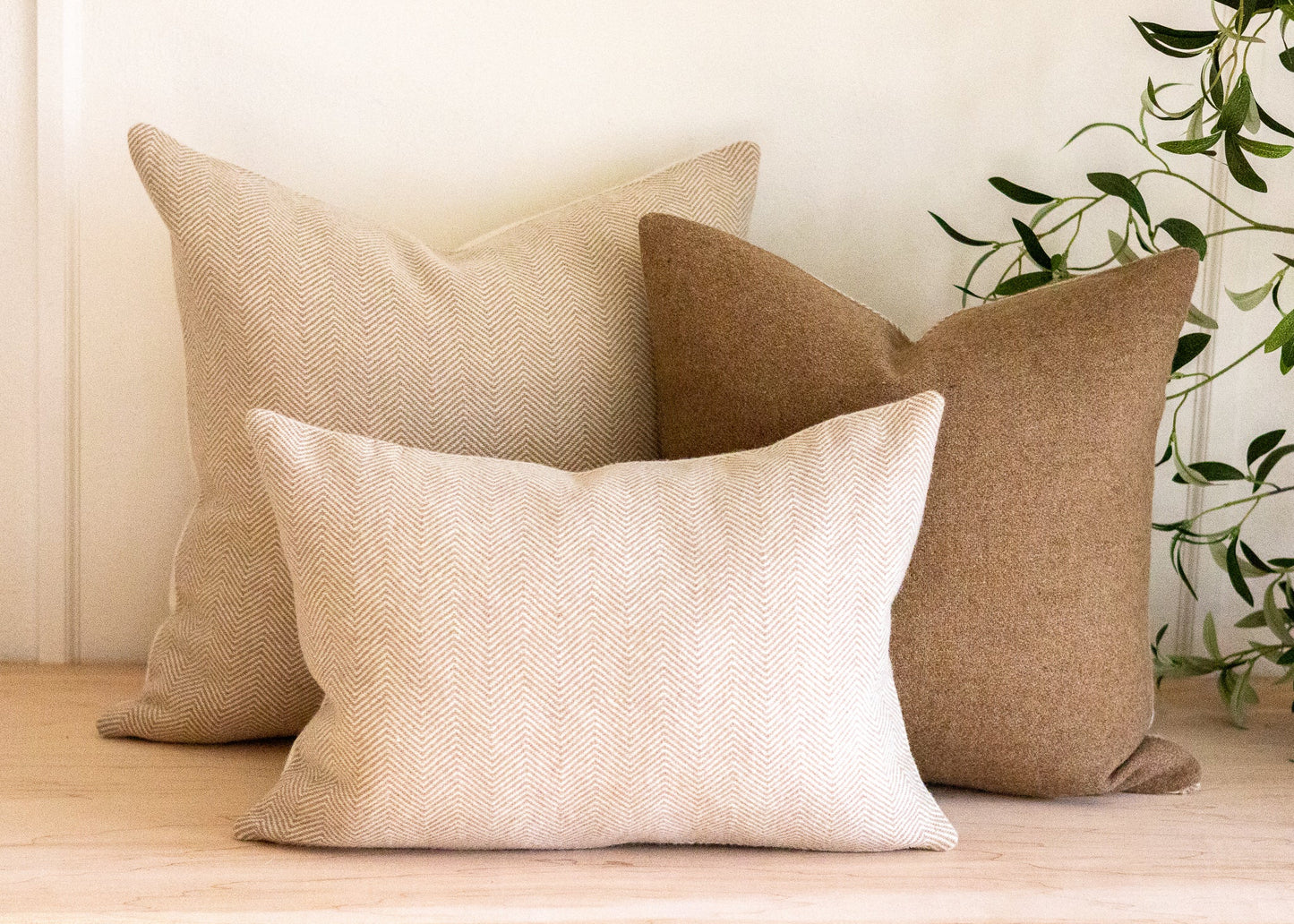Three beige and brown pillows on a wooden surface with a plant in the background.