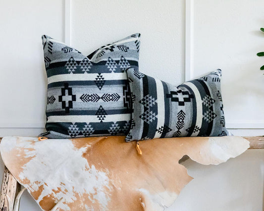 Two patterned pillows on a wooden bench with a white wall and plant in the background