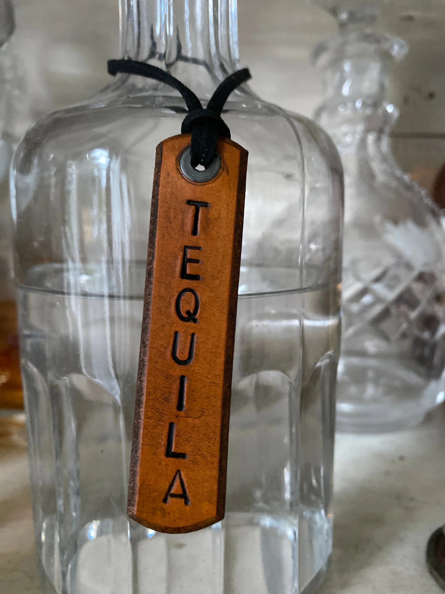 Clear glass bottle with a leather tag labeled 'Tequila' on a neutral background