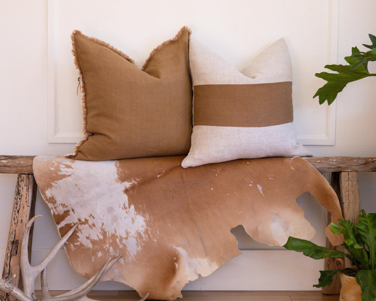Decorative pillows and cowhide rug on a wooden bench with plants and antlers.
