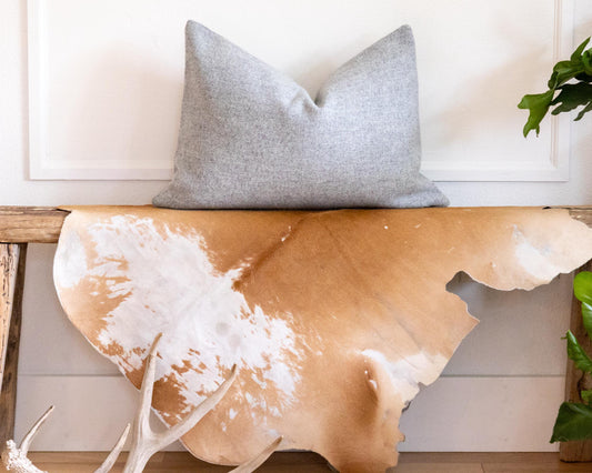 Gray pillow on a wooden bench with cowhide design, antlers, and plants in a room.