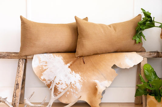 Two brown pillows on a wooden bench with a cowhide rug and antlers in the foreground.