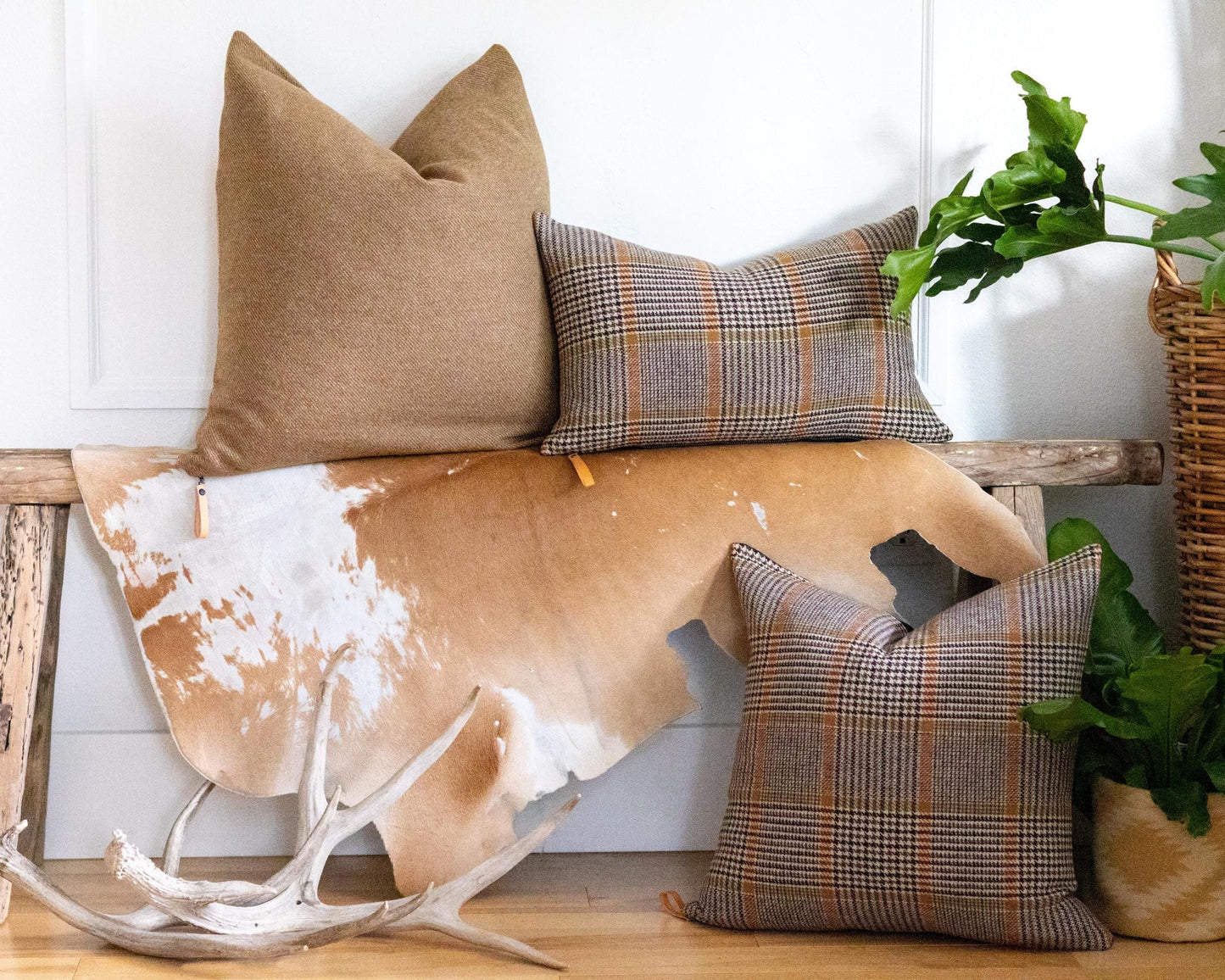 Decorative pillows on a wooden bench with a cowhide rug and antlers, against a white wall.