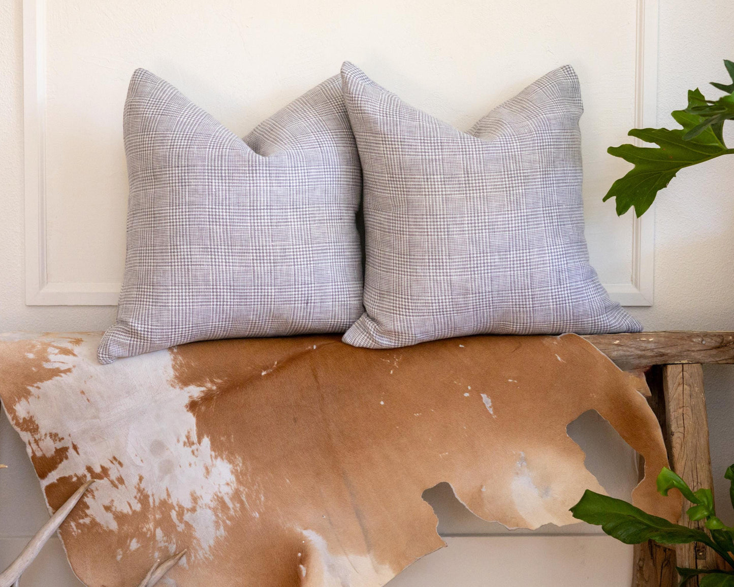 Two gray checkered pillows on a wooden bench with a cowhide rug and plants in the background.