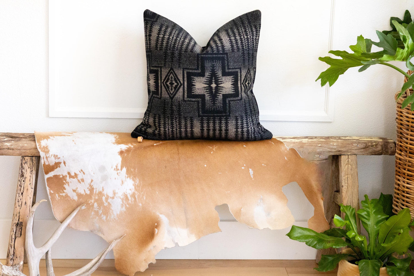 Decorative black and tan patterned pillow on a wooden bench with cowhide rug and plants