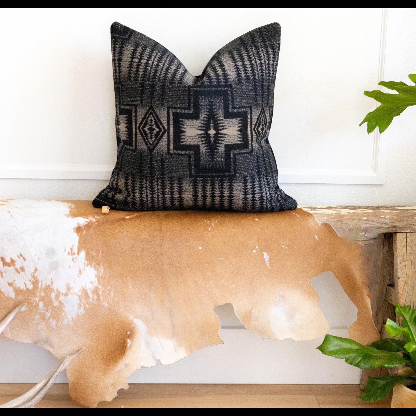 Decorative black and tan patterned pillow on a wooden bench with cowhide rug and plants