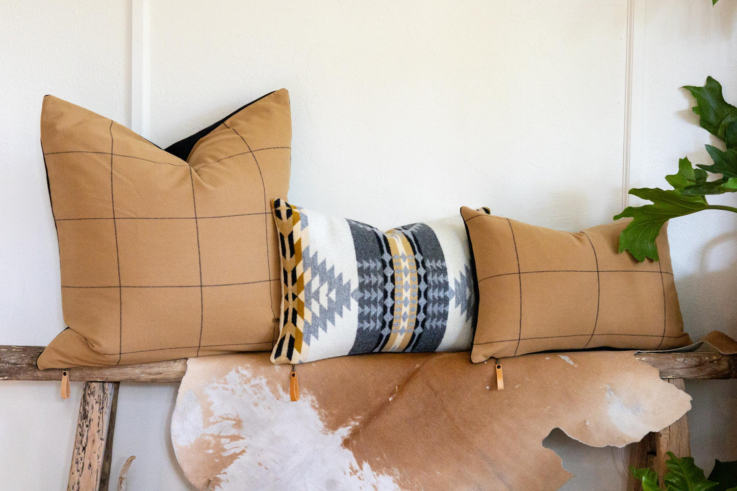 Three pillows on a wooden bench with a white wall and plant in the background