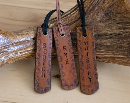 Three leather tags with embossed text 'SCOTCH', 'RYE', and 'WHISKEY' on a wooden surface.