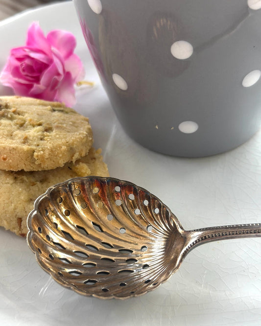 Silver-plated antique sugar sifter on a white surface with cookies and a polka dot mug in the background.