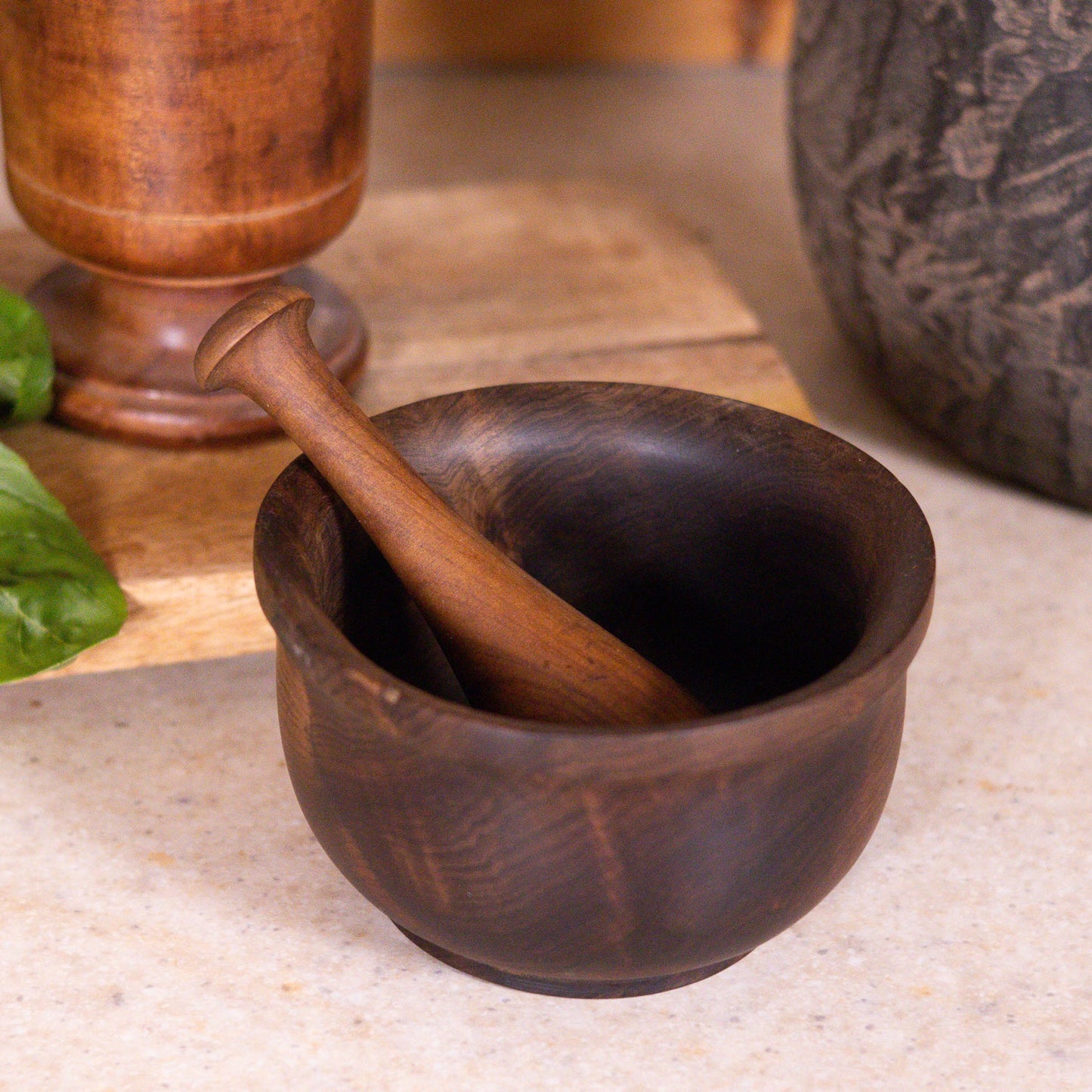 Wooden mortar and pestle on a stone surface with a wooden container and green leaves in the background.