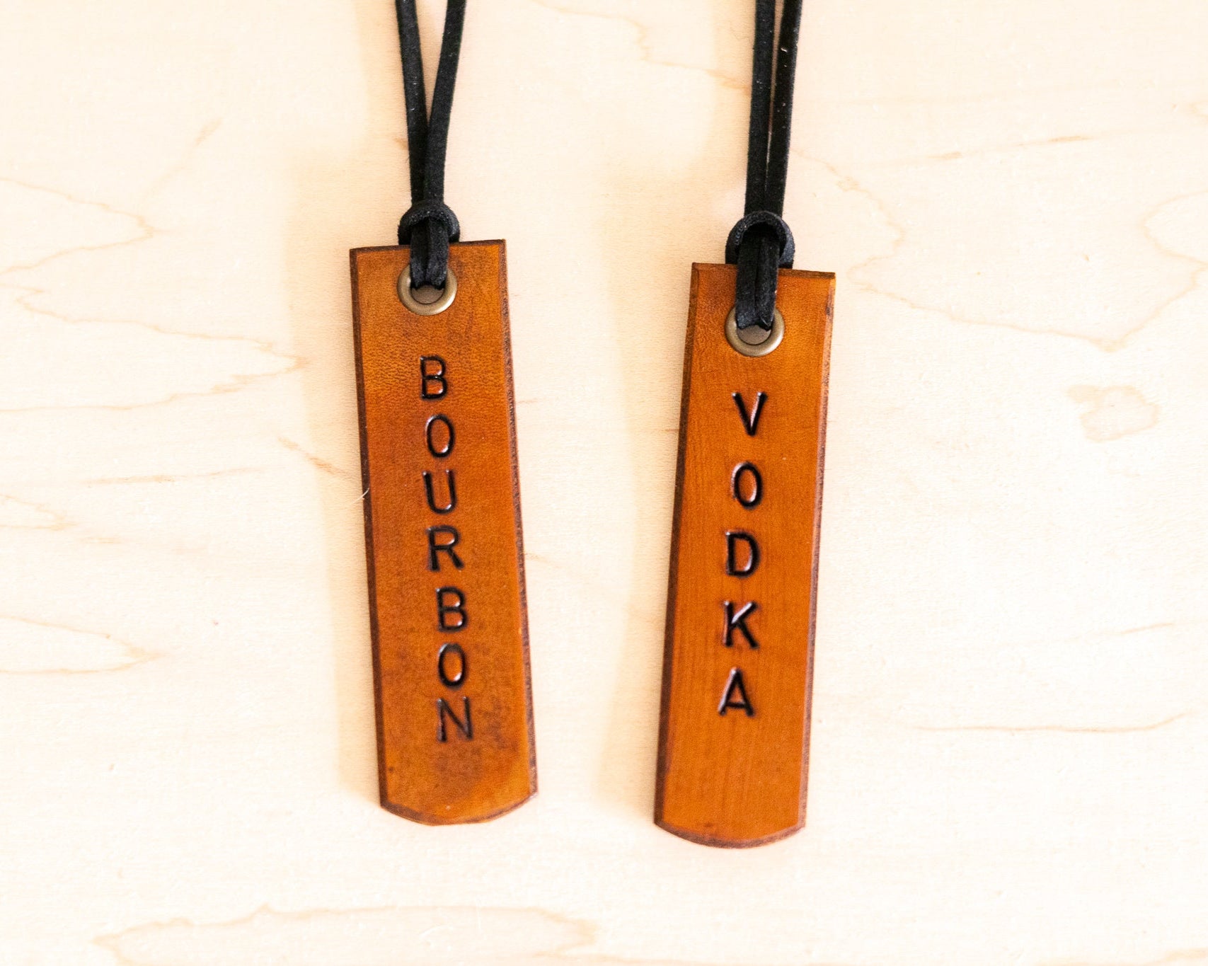 Two leather tags with 'Bourbon' and 'Vodka' engraved on them, hanging from leather strings.