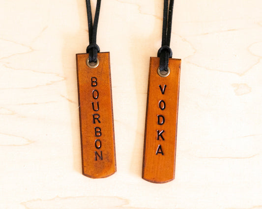 Two leather tags with 'Bourbon' and 'Vodka' engraved on them, hanging from leather strings.