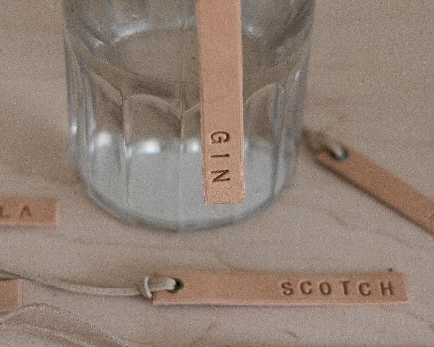 Clear Gin decanter bottle with leather tags labeled 'Gin', 'Scotch', and 'Rye' on a wooden surface.