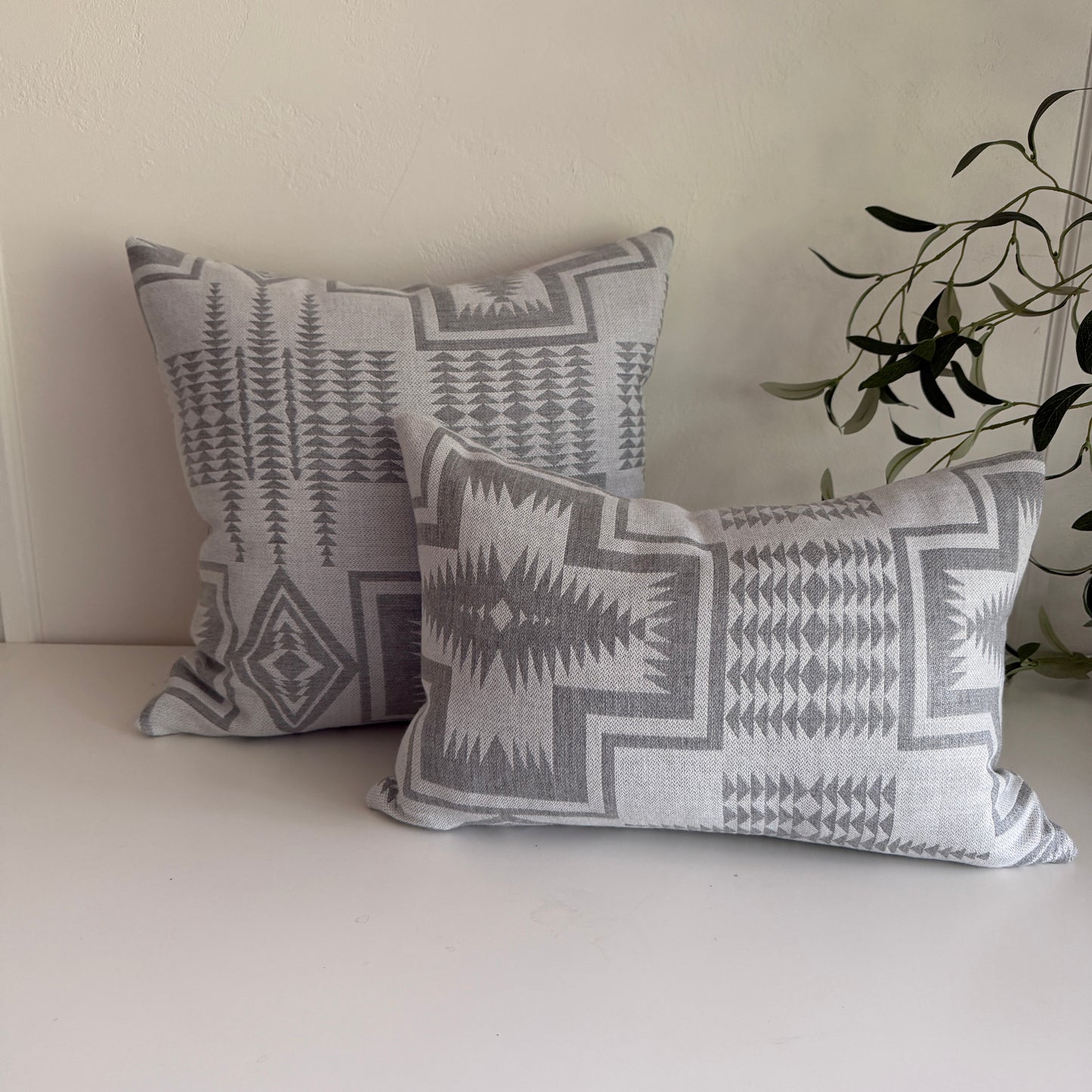Two gray patterned pillows on a white table with a plant in the corner.