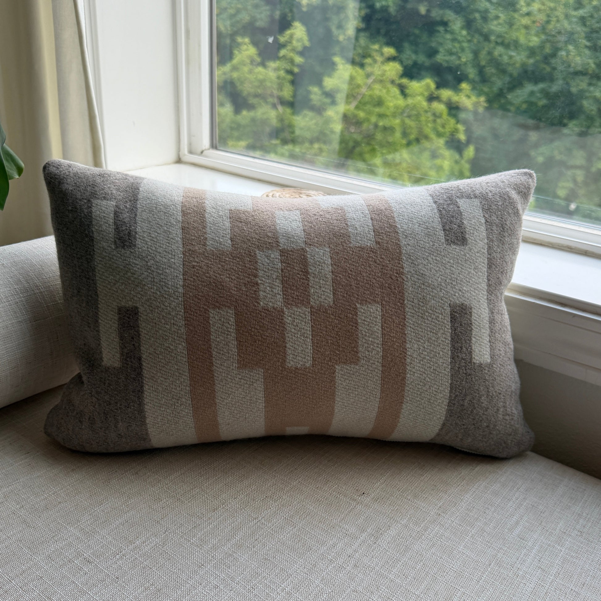 Decorative pillow with geometric pattern leaning against a light-colored wall next to a window with a view of greenery.