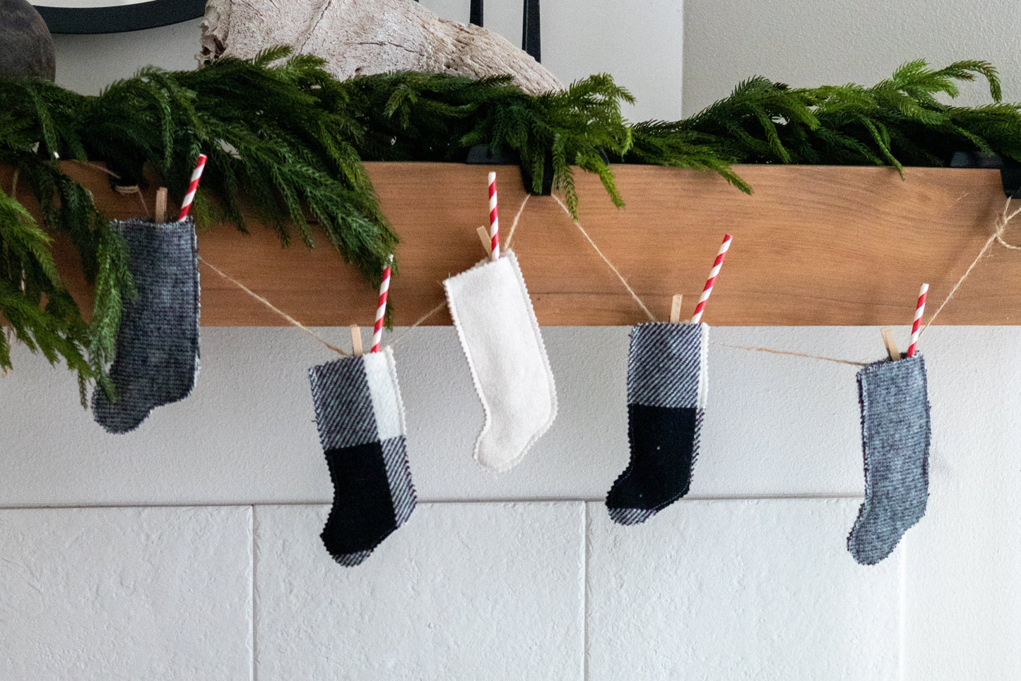 Miniature Christmas stockings hanging on a line with greenery above, against a white wall.