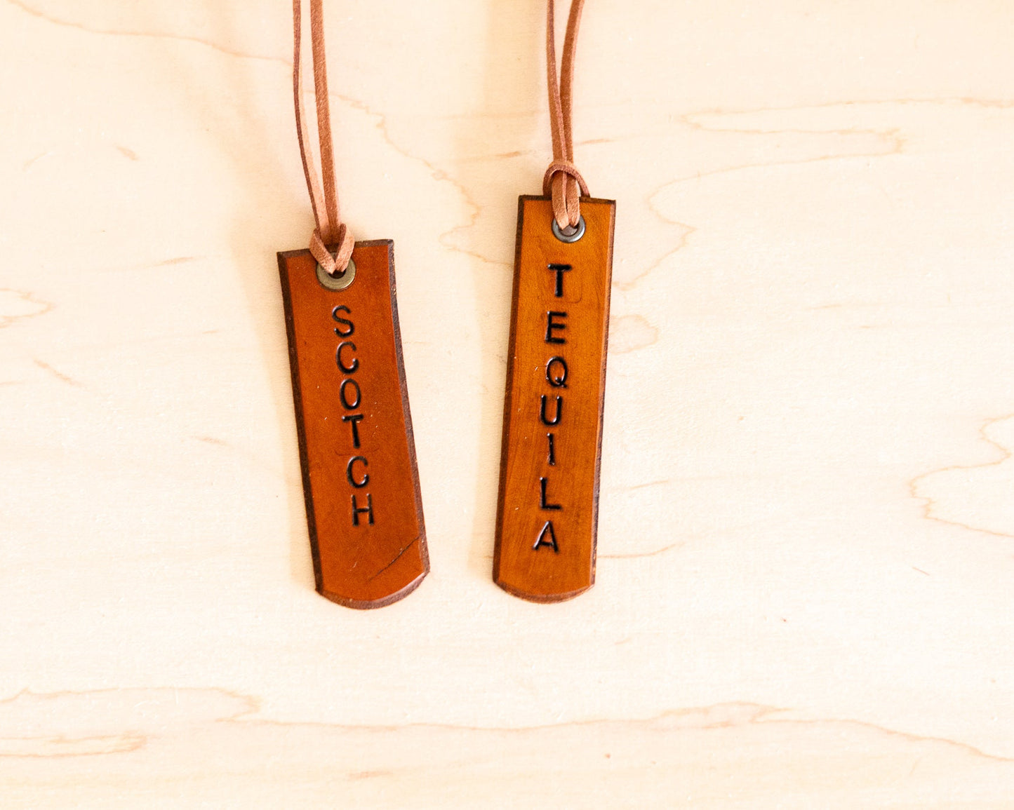 Two leather tags, one labeled 'Scotch' and the other 'Tequila', on a wooden background.
