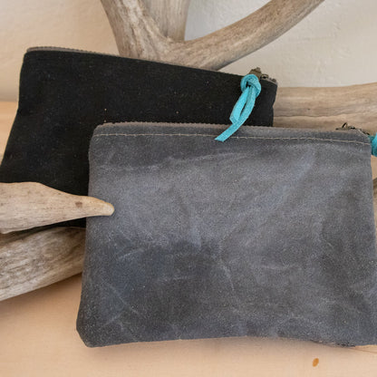 Two suede pouches, one black and one gray, with turquoise drawstrings, on a wooden surface with driftwood.