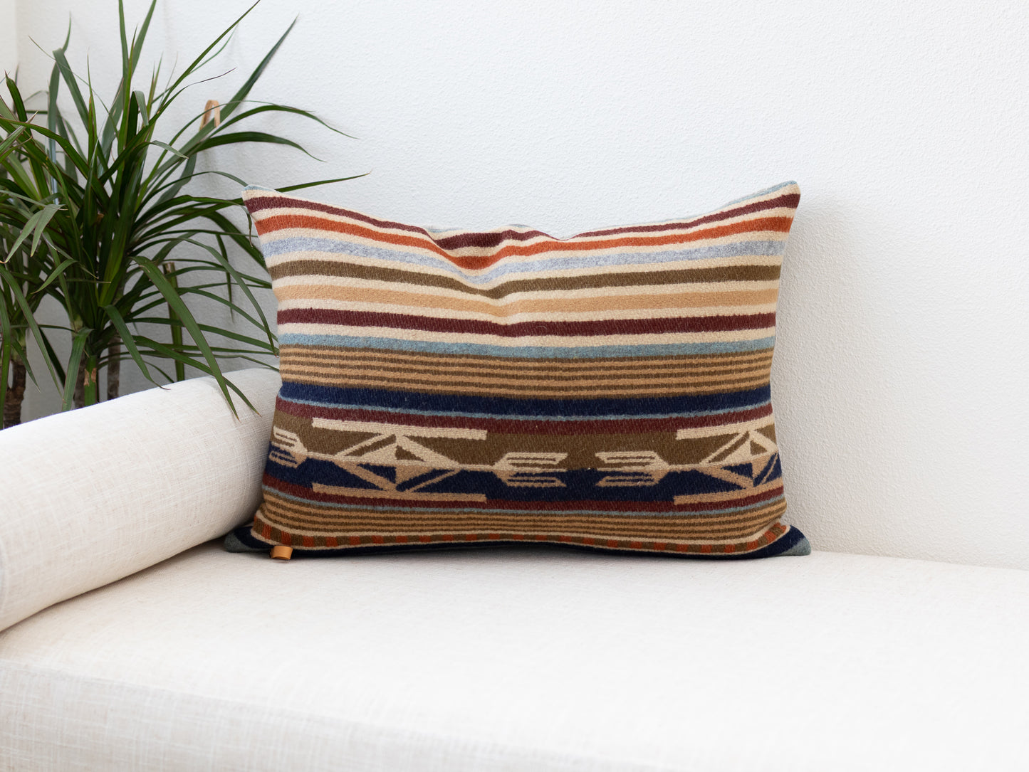 Striped wool lumbar pillow cover in rust, navy, and tan with a Southwestern geometric band, styled on a white sofa beside a potted plant.