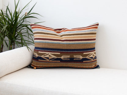 Striped wool lumbar pillow cover in rust, navy, and tan with a Southwestern geometric band, styled on a white sofa beside a potted plant.