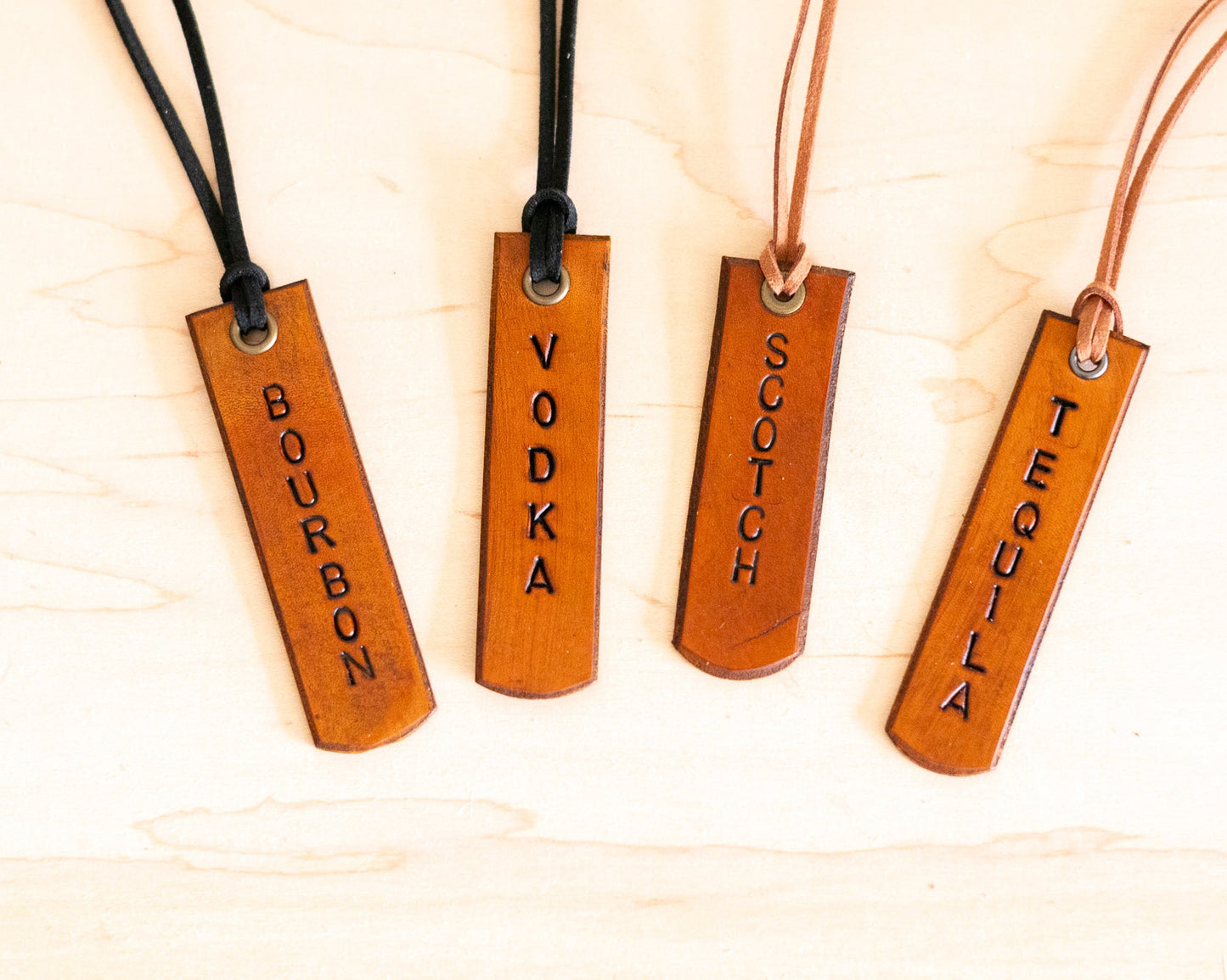 Four leather tags labeled 'Bourbon', 'Vodka', 'Scotch', and 'Tequila' on a light wooden background.
