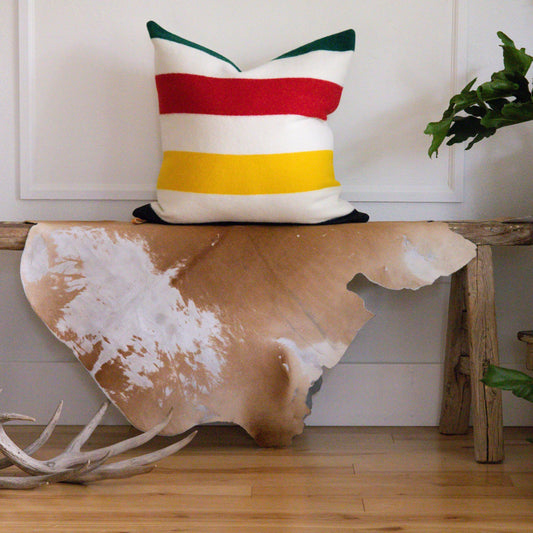 Striped pillow on a wooden bench with a cowhide rug and antlers in a room.