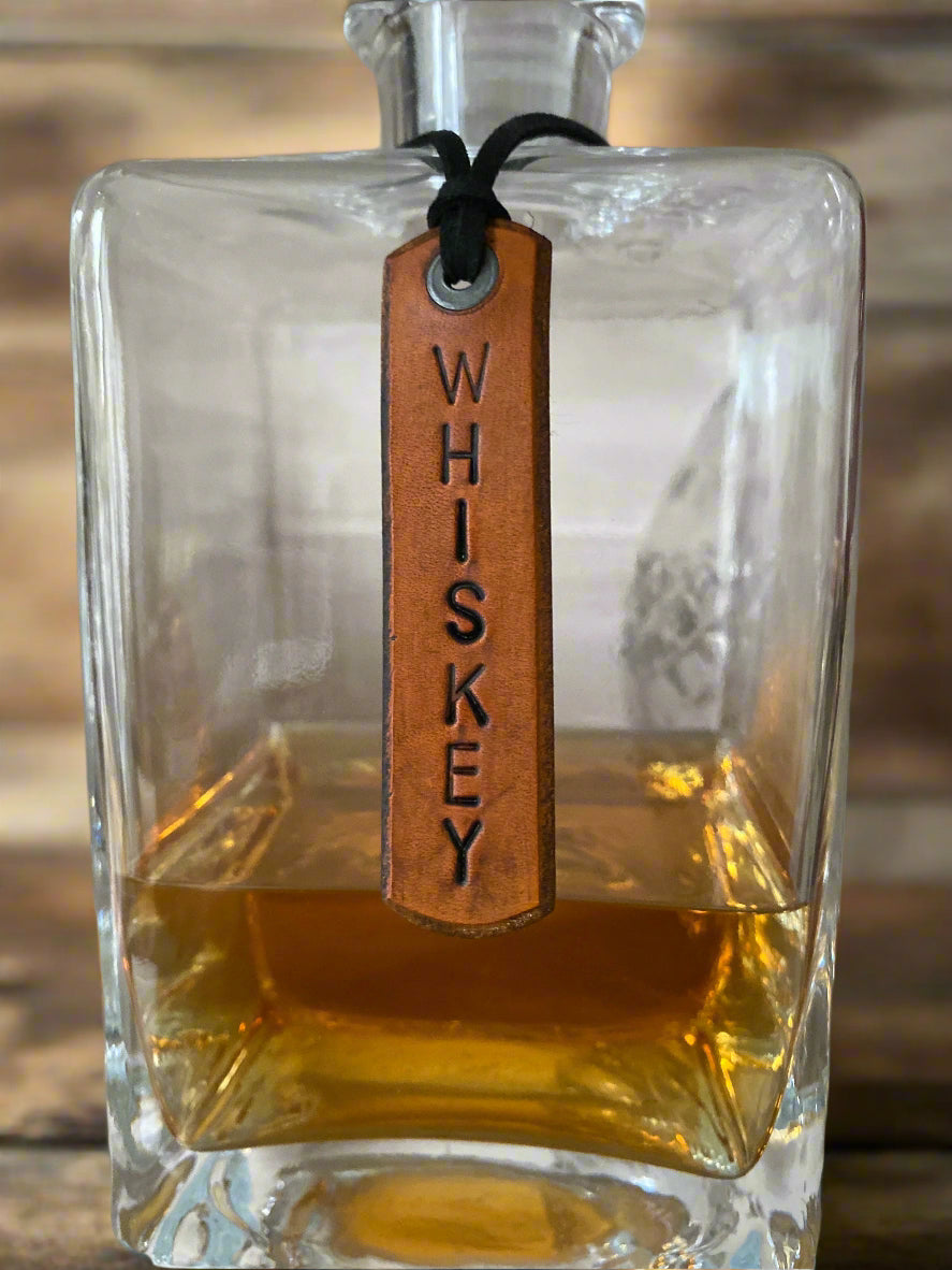Glass bottle of whiskey with a leather tag labeled 'Whiskey' on a wooden surface.
