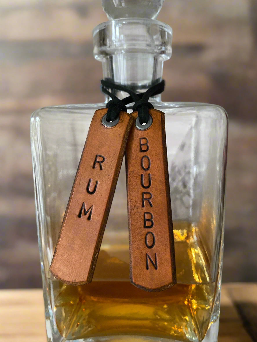 Glass decanter with 'Rum' and 'Bourbon' leather tags on a wooden surface.