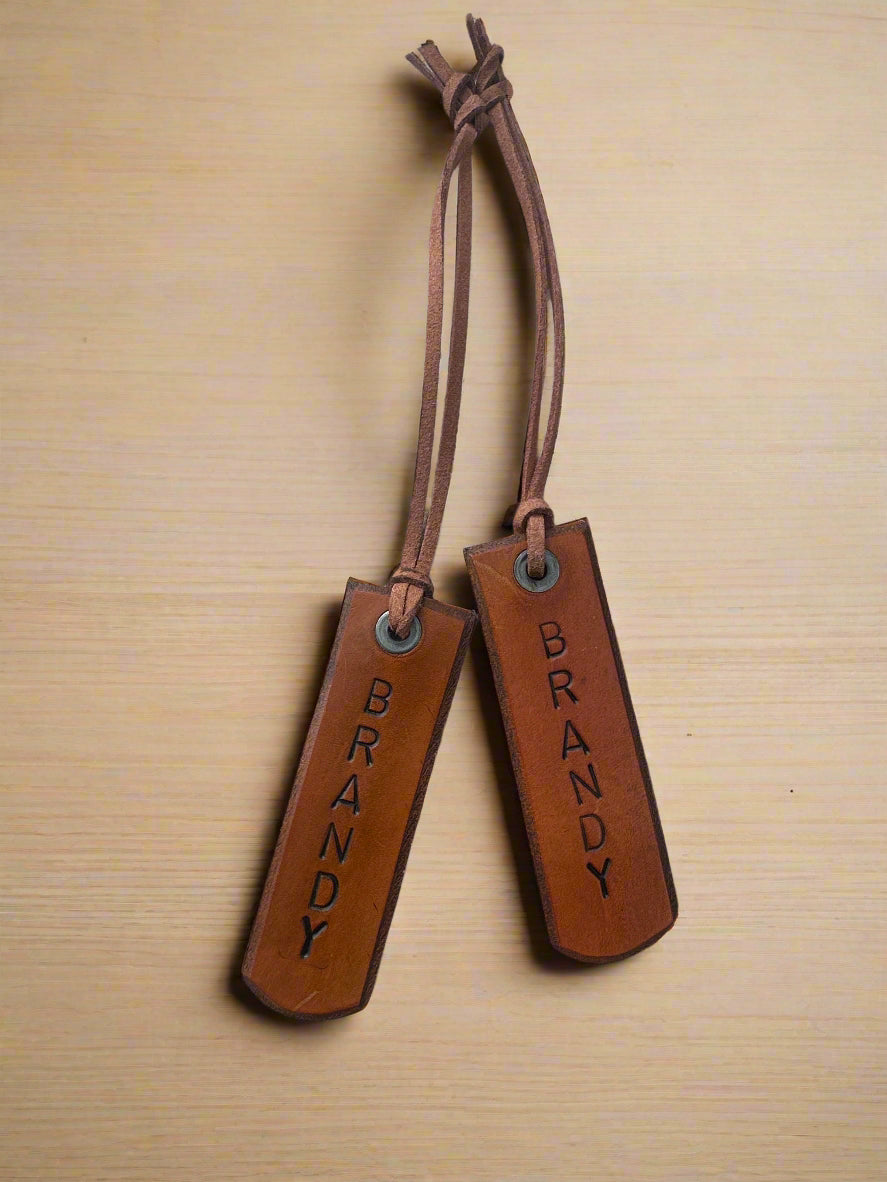 Two brown leather luggage tags with 'BRANDY' engraved on a wooden surface.
