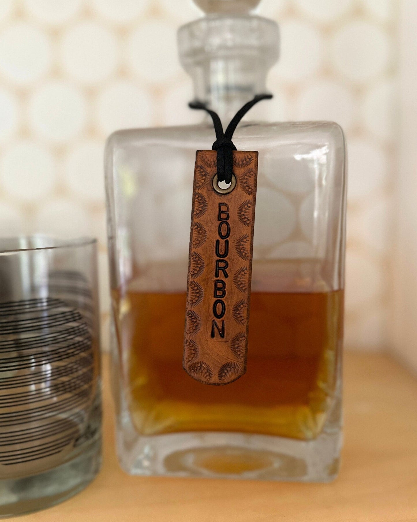 Glass decanter with a leather tag labeled 'Bourbon' next to a glass on a wooden surface.