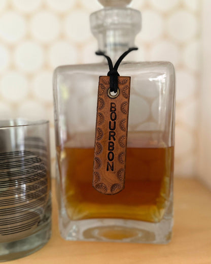 Glass decanter with a leather tag labeled 'Bourbon' next to a glass on a wooden surface.