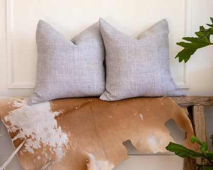 Two gray checkered pillows on a wooden bench with a cowhide rug and plants in the background.