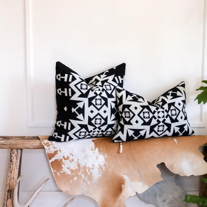 Two black and white patterned pillows on a wooden bench with a white wall background.