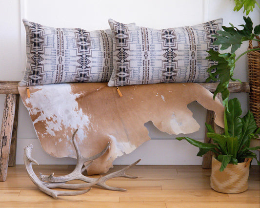 Decorative Pendleton® pillows with geometric patterns on a cowhide rug with antlers, against a white wall.